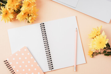 Desktop with notebook, laptop computer, pencil, bouquet of yellow flowers on beige background. Top view, flat lay