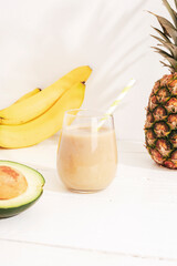 Fototapeta na wymiar Banana, pineapple and avocado smoothie on white wooden table in sunlight. Minimal detox diet concept, summer vitamin drink