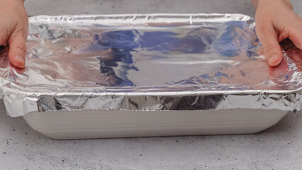 Baking dish covered with aluminum foil close up on the table, front view, woman hands. Spinach lasagna recipe.