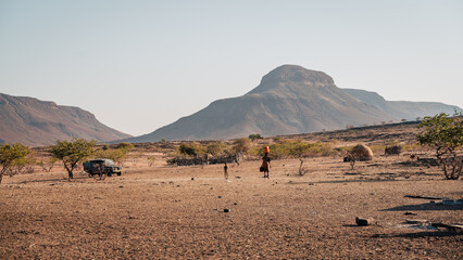 Blick auf ein Himba-Dorf in der kargen Landschaft des Kaokoveld - Im Mittelpunkt des Bildes sieht man eine Frau mit einem Wasserkanister auf dem Kopf (Kunene, Namibia)