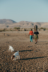 Blick in ein Himba Dorf - Eine Frau, die ein Gefäß auf dem Kopf trägt, läuft mit ihrer...