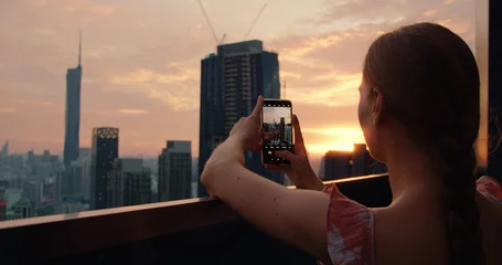 Fotobehang Woman on roof of skyscraper shoots a city landscape on a mobile phone during sunset. Rear view of tourist girl in twiligh on vacation. Kuala Lumpur capital of Malaysia. © vidoc