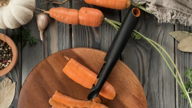 Peeling ripe orange carrot with vegetable peeler and knife. Stop motion, flat lay. Fresh unpeeled carrots. Skinning, cutting peel on kitchen wooden board. Prepare ingredients for cooking, recipe.