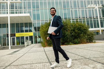 Smiling indian businessman in suit and glasses with laptop near office building