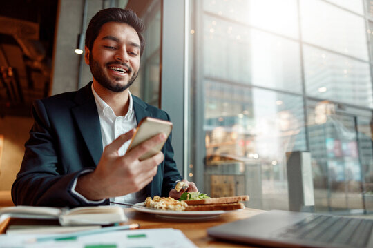 Smiling Indian businessman wearing suit is using phone during lunch time in cafe. Blurred background