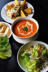 Lunch set made of tomato cream soup, salad, chicken with potato, lemonade on black background