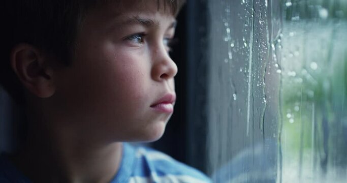 Sad little boy with mental issues, looking alone and bored while watching the rain from a window. Ptsd, abuse and trauma victim stuck in a bad, toxic environment. Orphan feeling lonely and depressed