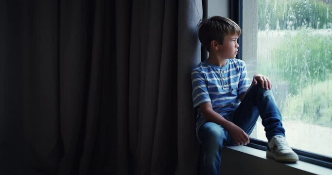 Sad, depressed and upset young child sitting by the window and watching the rain on a cold, wet and rainy winter day. A lonely little boy at home and unhappy about not being able go play with friends