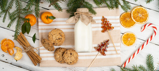 milk and cookies on the table with spruce branches, christmas treats for Santa Claus, tangerines, cinnamon, christmas traditions, flat lay, banner