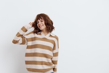 a beautiful, pleasant woman stands in a stylish striped sweater with her hands raised behind her head on a light background, closing her eyes and smiling broadly