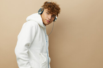 a cute, attractive man stands on a beige background in a white hoodie and listens to music with headphones. Horizontal photo
