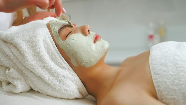Face clay mask concept. Indoor closeup portrait of young positive Asian girl lying down with sage green clay mask spread on her face by professional cosmetician. High quality 4k footage