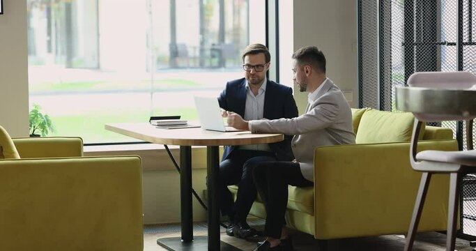 Male sales manager makes commercial offer to client during meeting in modern office, two businessmen talking sitting on sofa at table with laptop at workplace, share ideas, discuss online presentation