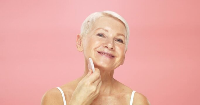 Beauty portrait of senior woman using gua sha massager for perfect skin on cheeks of her face, isolated on pink background