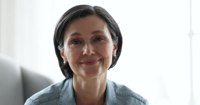 Close up attractive brunette middle-aged 55s female smile staring at camera seated indoor. Portrait of happy retiree woman spend time alone at home, having pleasant appearance, looks good, enjoy rest
