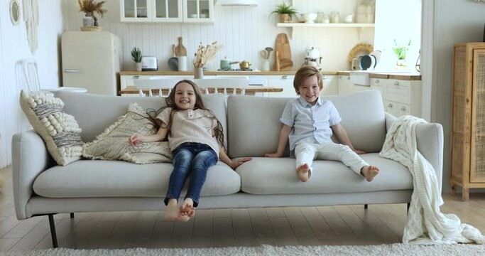 Two happy little 6s kids jumping on couch relaxing together in studio flat. Preschoolers cousins, friends or siblings have fun at home, flopped down on soft sofa enjoy rest and playtime at cozy home