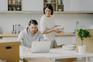 Young brunette woman unpacks boxes with personal belongings, places plates in cupboard, looks attentively at laptop computer where husband works. Family couple relocate in new modern apartment