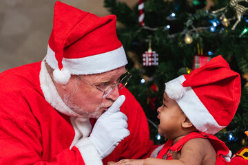 Santa claus is hushing the crying little toddler baby with the christmas tree on the back