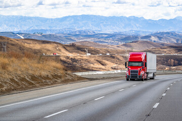 Bright red big rig bonnet semi truck transporting cargo in dry van semi trailer slowly climbing uphill on the mountain highway road