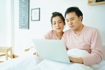 Affectionate Asian lovely couple laying on a bed in bedroom and browsing the internet on laptop or notebook computer together in weekend.