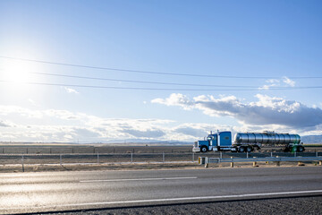Naklejka premium Classic powerful blue big rig semi truck with chrome accessories transporting liquid cargo in stainless steel tank semi trailer driving on the straight highway road between fields