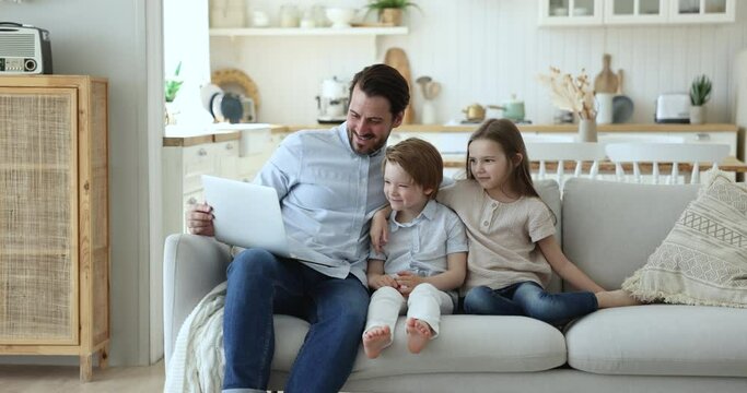 Young father and preschoolers daughter and son spend weekend together sitting on couch at home using modern laptop, watching movie, cartoons or vlog on internet. Modern technology, habit, lifestyle