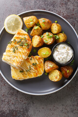 Healthy food serving grilled fish cod with baked potatoes, sauce and lemon close-up in a plate on the table. Vertical top view from above