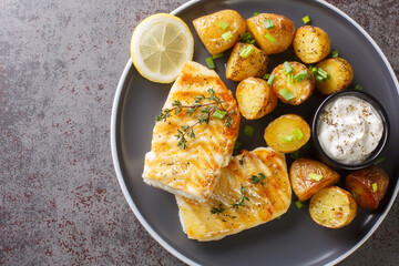 Portion of grilled cod fish with baked jacket potatoes, creamy sauce and lemon close-up in a plate on the table. Horizontal top view from above