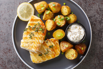 Close-up dish of grilled codfish with baked jacket potatoes, creamy sauce and lemon on the table. Horizontal top view from above