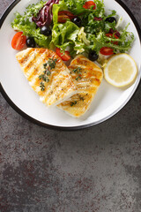 Healthy food serving grilled fish cod with fresh vegetable salad and leaf lettuce mix and lemon close-up in a plate on the table. Vertical top view from above