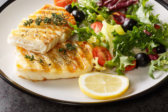 Delicious healthy grilled cod fish with fresh salad of tomatoes, olives, lettuce mix and lemon close-up in a plate on the table. Horizontal