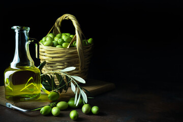 Glass jar of olive oil on wooden cutting board and a wicker basket with fresh green olives on black background. Copy space on right side. Dark low key photo. Blurred background. Selective focus. - Powered by Adobe