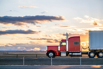 Side view of red big rig classic semi truck transporting cargo in dry van semi trailer moving on...