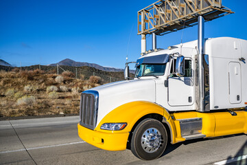 Two colors white and yellow big rig semi truck tractor with extended cab for truck driver rest transporting cargo running on the wide highway road with information board frame