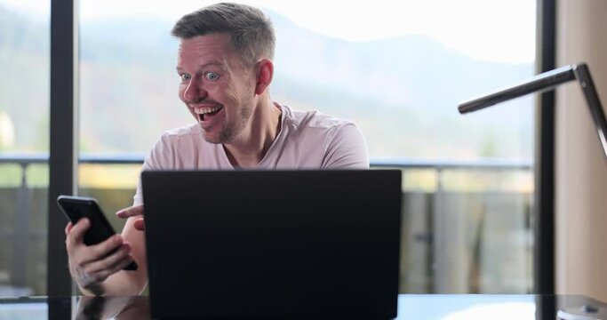 Man sits at table, reads email on smartphone and on laptop makes yes gesture and feels happy