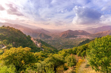 Obraz premium beautiful landscape with amazing view from highland with green slopes and bushes to a valley with town, road and nice mountains and scenic cloudy sunset on background