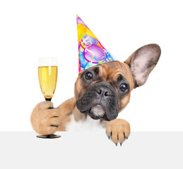 French bulldog puppy wearing party cap looks above empty white banner and holds glass of champagne. isolated on white background