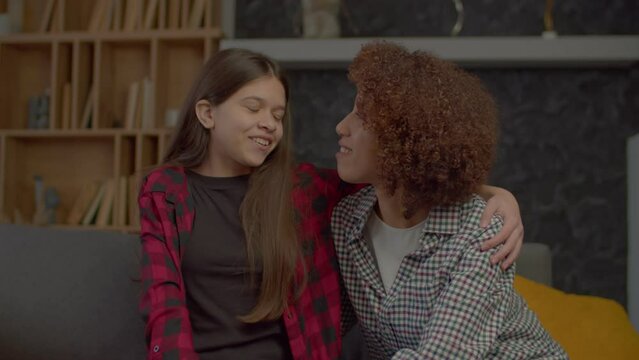 Affectionate beautiful African American mother and lovely multiracial teenager daughter embracing on sofa , talking and bonding, expressing happiness, love and strong relationship indoors.