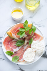 Slices of prosciutto ham with mozzarella, cantaloupe melon and fresh green basil, vertical shot on a white marble background