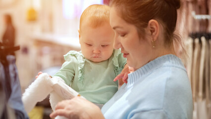 Baby girl with mother choose clothes. Child with amused and puzzled expression enjoys shopping with parent closeup, sunlight