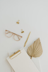 Flatlay of clipboard with blank copy space paper sheet. Tan fan leaf, glasses, pen, clips on white...