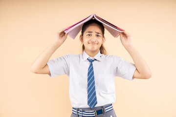 Unhappy Indian student schoolgirl wearing school uniform holding books on head standing against beige background, Education, Mental health, Stress. 