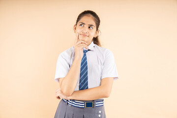 Fototapeta Modern Indian student schoolgirl wearing school uniform thinking expression standing isolated on beige background, Studio shot, closeup, Education concept. obraz