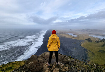 Adventurer in yellow jacket overlooks scenic coastline reynisfjara black beach VIK from a cliff on Dyrholaey Lighthouse in Golden Circle in Iceland