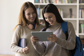 Cheerful Latin student girl showing learning online presentation on tablet to classmate, holding...