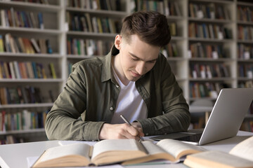 Positive busy hardworking student guy writing draft, notes, article, essay, webinar summary, studying in college library, working on research at desk with open books, laptop computer