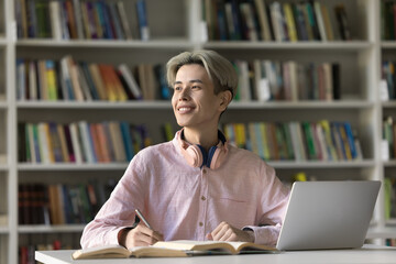 Joyful teenage Asian student doing homework in college library, writing notes at laptop and open...