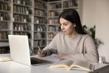 Positive beautiful Hispanic teenage student girl watching learning webinar, online class, video lesson on laptop, writing notes, summary, studying in university library, sitting at open book, smiling