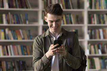 Happy handsome student guy using learning application for study on smartphone, reading message, typing, chatting, watching content in college library, smiling, laughing, holding cellphone