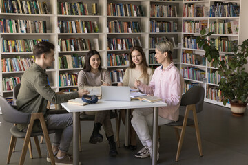Happy multiethnic team of college students studying in university library, talking, laughing, discussing homework task, enjoying teamwork, education process, sitting at open books and laptop